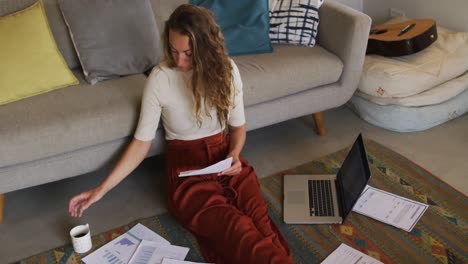 mujer caucásica trabajando en la sala de estar, sentada en el suelo con una computadora portátil y papeleo, bebiendo café