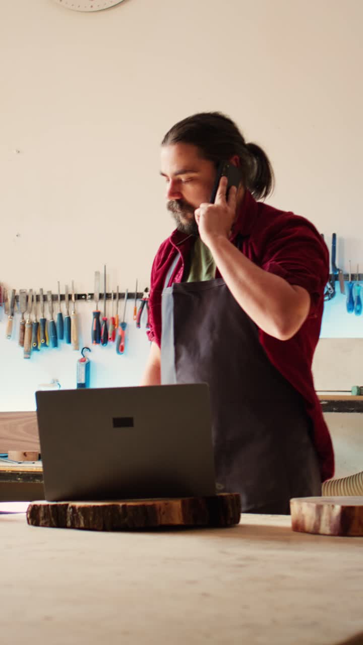 Artesão falando ao telefone enquanto trabalha no laptop na oficina