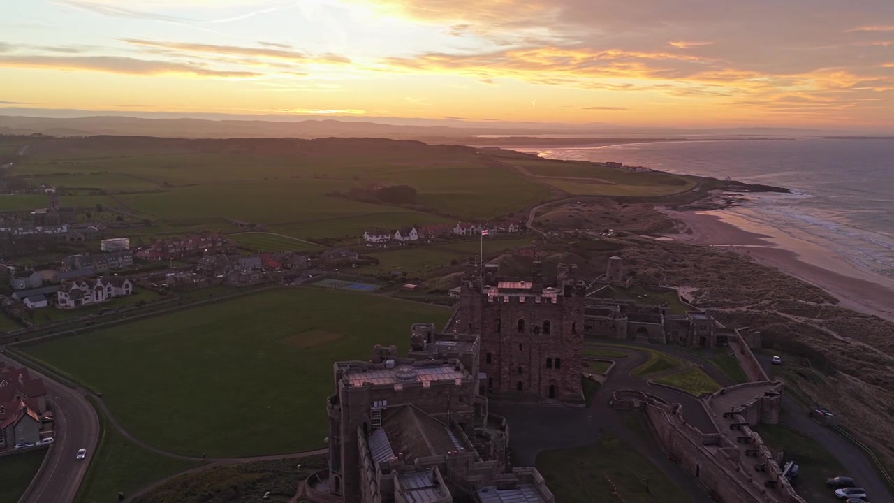 Majestetisk luftopptak som fanger Bamburgh Castle i Northumberland, England. Dronen sirkler sakte rundt den historiske festningen under en vakker solnedgang.