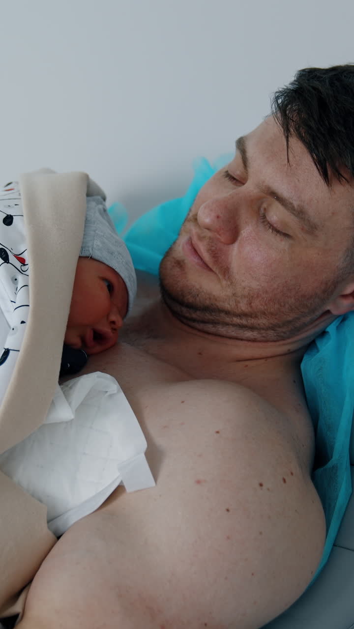 Muscular Caucasian man with bare chest holds his newborn. Skin to skin contact for a C-section born child in hospital. Vertical video