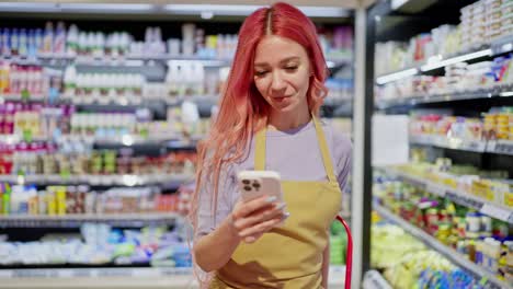 a girl with pink hair in a yellow apron scrolls through social networks during a break at work among dairy products in a supermarket