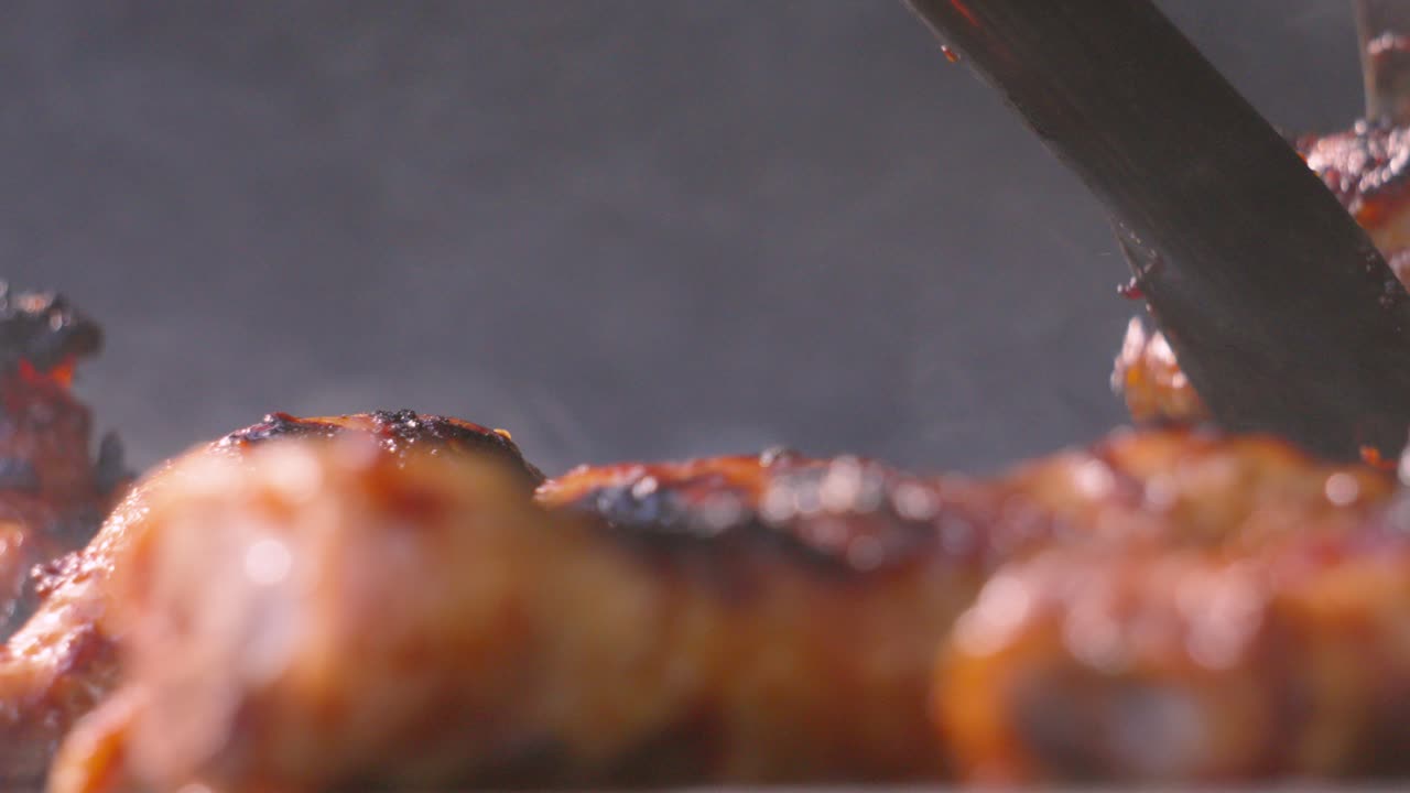 Level View of BBQ with Smoke Rising from Hot Charcoal Below as Tongs Come Down and Pick Up Pieces of Chicken to Serve. Summer Outdoor Barbecue Food with Whole Pieces of Delicious Meat.