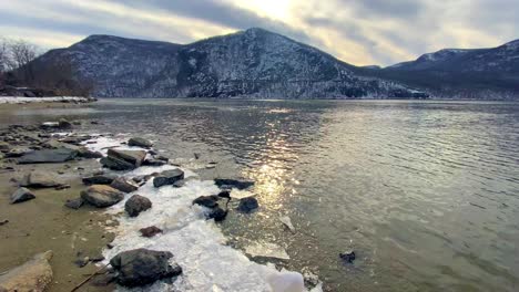 Beautiful-snow-covered-mountains-over-a-wide,-flowing-river-with-beautiful-overcast-skies-during-winter-in-the-Appalachian-mountains-with-an-icy-shore