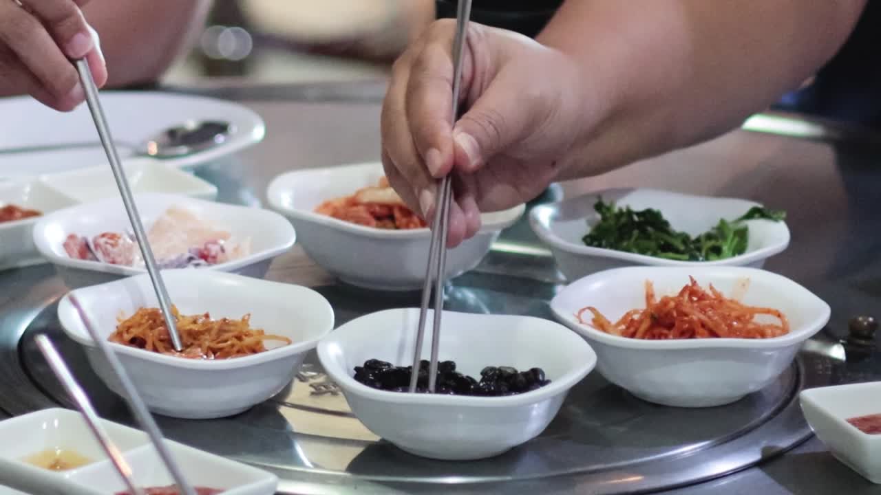Close-up of hands using chopsticks to share various Korean side dishes on a round table.