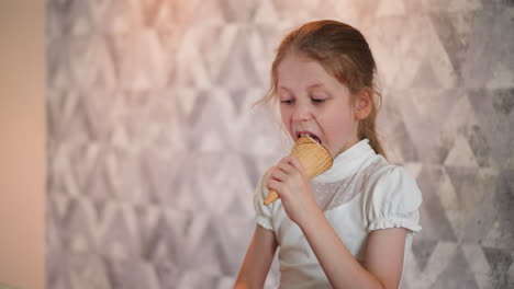 young girl in white short-sleeve top stands in front of textured patterned wall enjoying cone of ice cream while casually engaging in relaxed indoor activity
