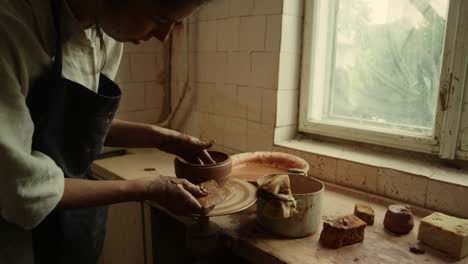 Focused-master-making-new-clay-product-in-pottery.-Woman-scraping-line-on-pot