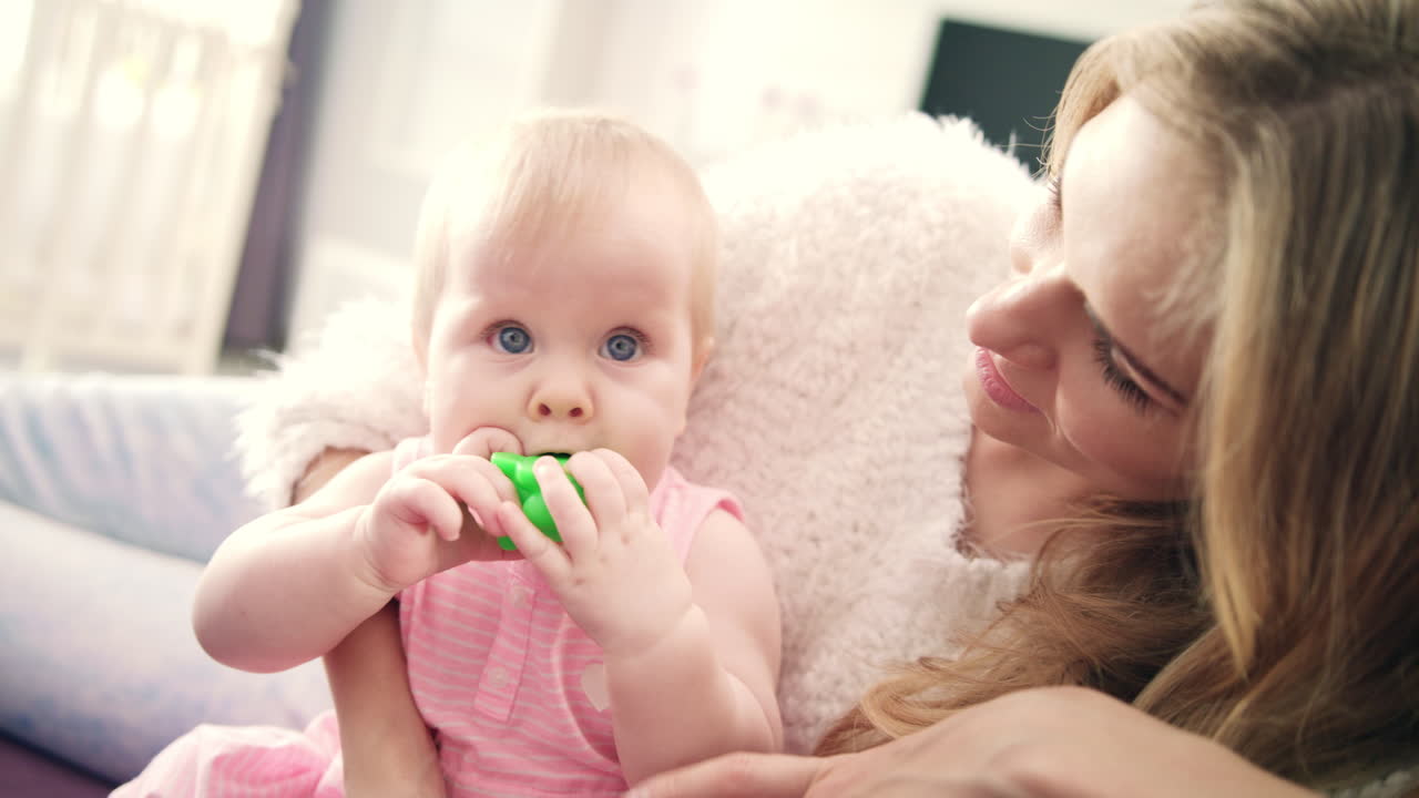 Beautiful baby girl sitting in mother embrace. Toddler girl with toy in mouth