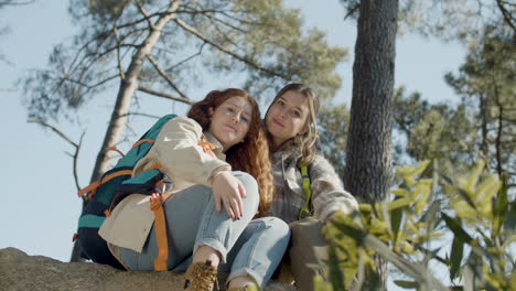 two young female hikers sitting on a stone, hugging and looking at the camera