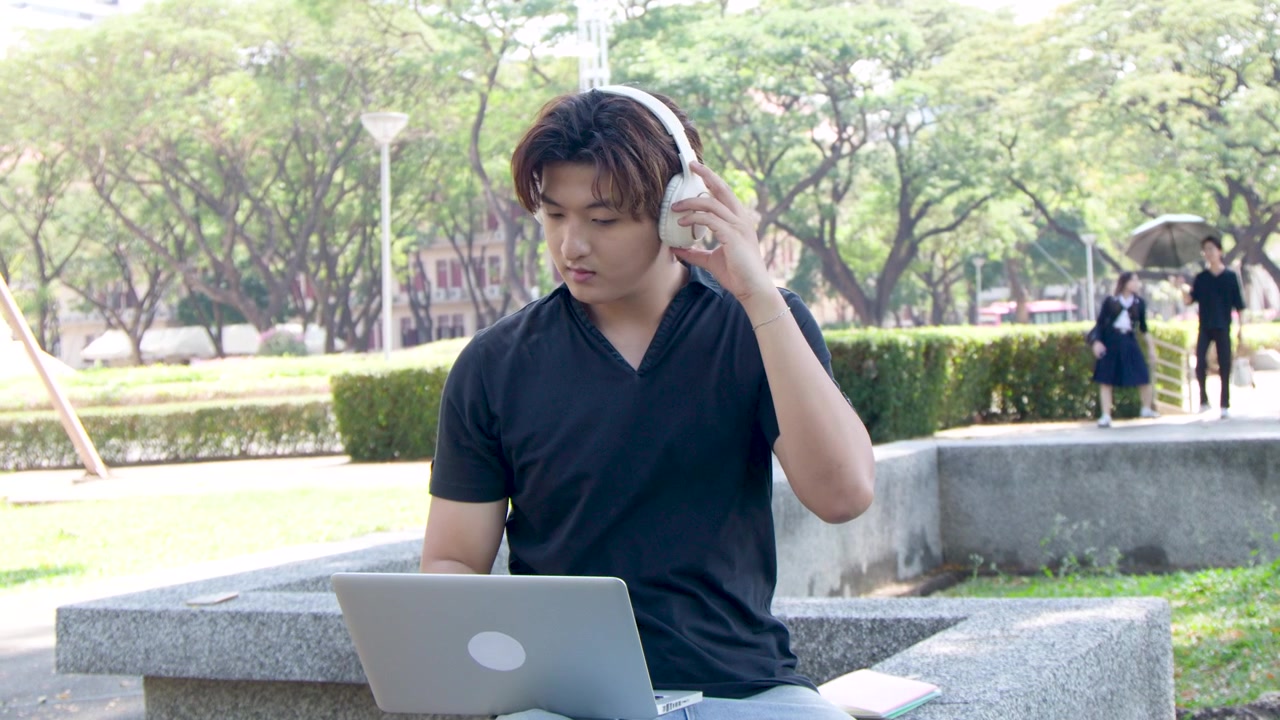 ชายหนุ่มชาวไทยนั่งอยู่กลางแจ้งที่สวนสาธารณะของมหาวิทยาลัย กำลังใช้แล็ปท็อปและหูฟัง