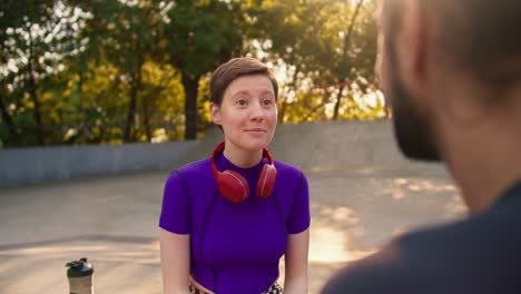 Un-Chico-Moreno-Con-Una-Camiseta-Gris-Se-Comunica-Con-Una-Chica-Con-Un-Corte-De-Pelo-Corto-Con-Un-Top-Morado-Y-Auriculares-Rojos-En-Un-Parque-De-Patinaje-En-Verano