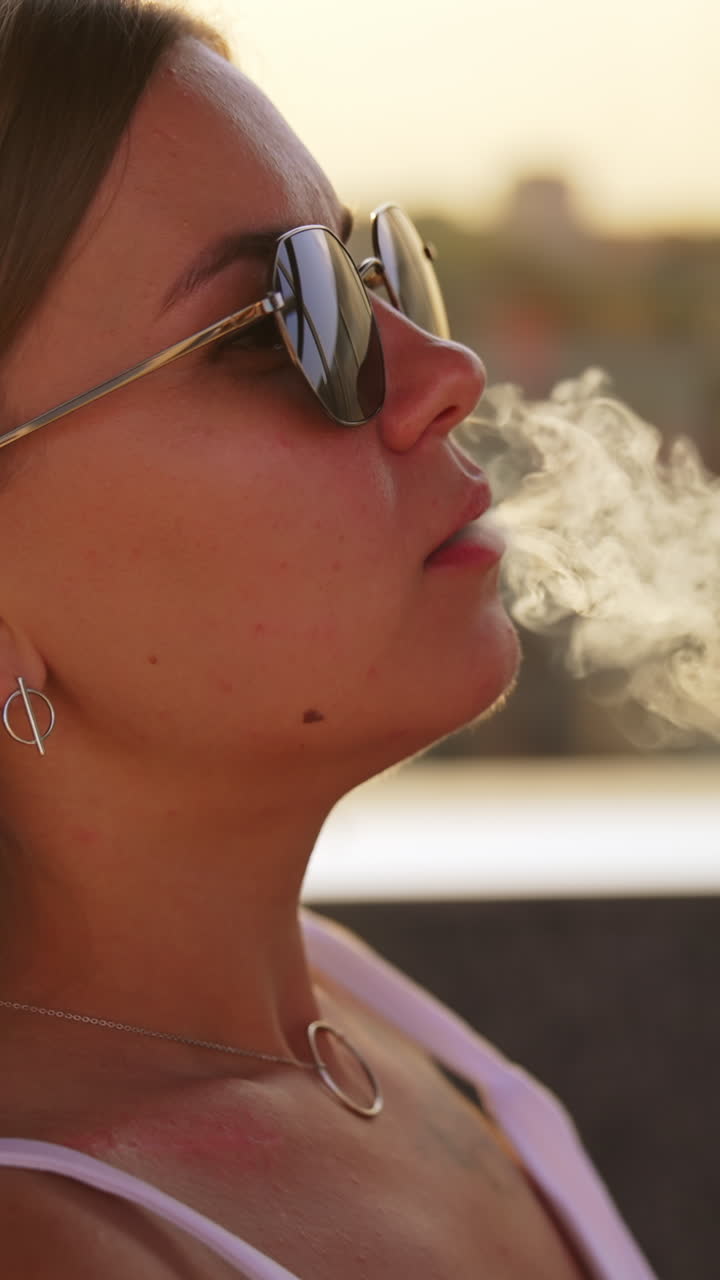 Smoking hookah in a café. Young long-haired woman breathes in and out thick white smoke. Close up. Blurred backdrop. Vertical video