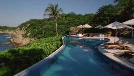 Aerial-view-over-a-luxurious-blue-swimming-pool-in-motion-at-a-hotel-in-Huatulco,-surrounded-by-swaying-palm-trees,-moving-backward