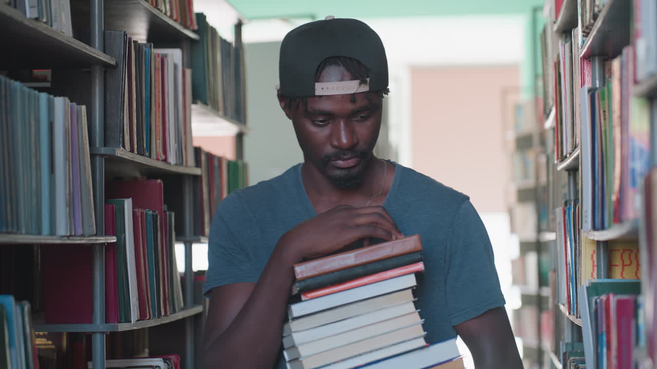 Man walking along library bookshelf carrying stack of books while looking around thoughtfully, surrounded by colorful book spines in well-lit aisle, wearing cap and casual shirt
