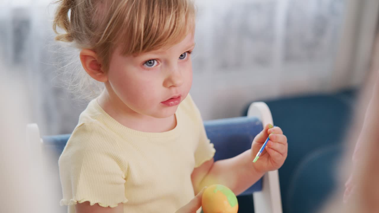 Young girl paints Easter egg with brush. Mother smiles beside her. Focus on child's concentrated expression. Soft, natural light enhances warm, joyful atmosphere. Ideal for family, spring, holiday