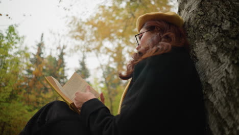 lower angle view of woman in yellow beret with red hair cascading down her shoulder as she carefully opens a page of her book, immersed in deep thought