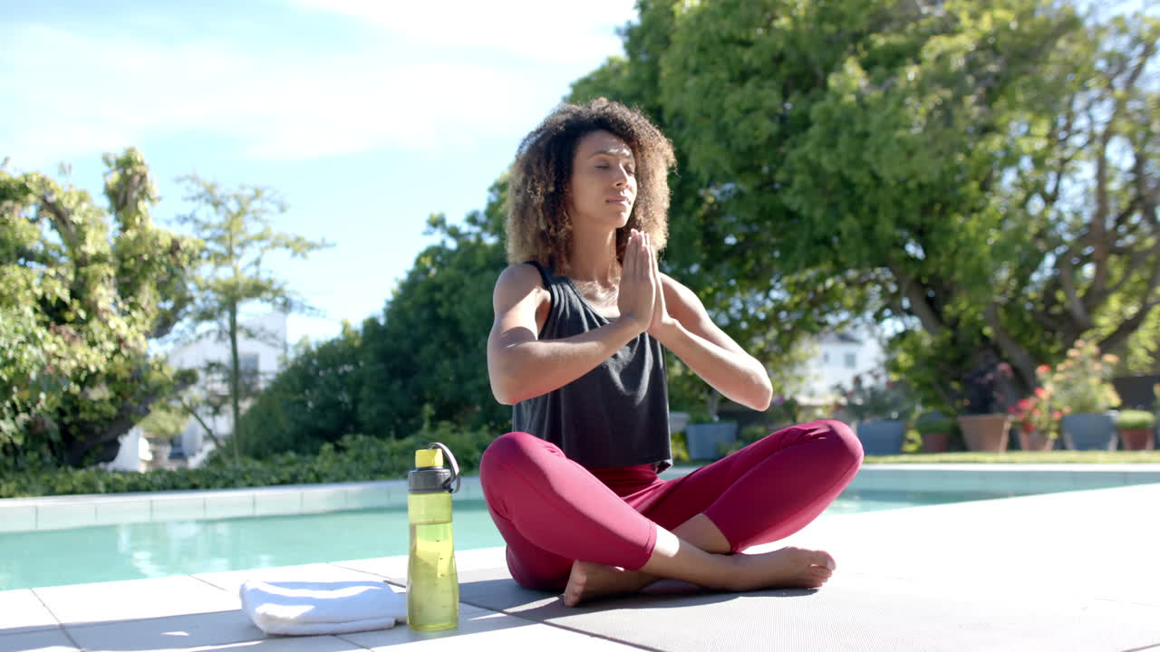 feliz mujer biracial practicando meditación de yoga sentada junto a la piscina en un jardín soleado, cámara lenta