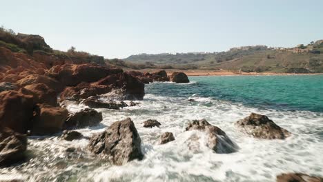 Flying-fast-and-low-above-seashore-with-beautiful-turquoise-waters-of-the-Mediterranean-Sea-splashing-on-the-red-coloured-rocks