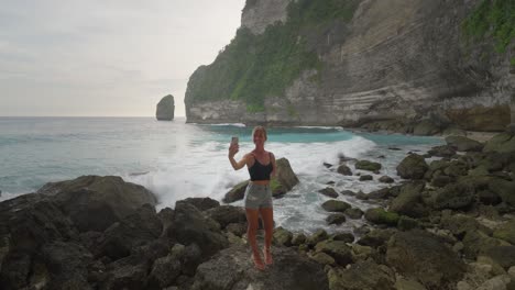 Fit-female-vlogger-posing-and-smiling-at-phone-camera-while-waves-crash-in-background