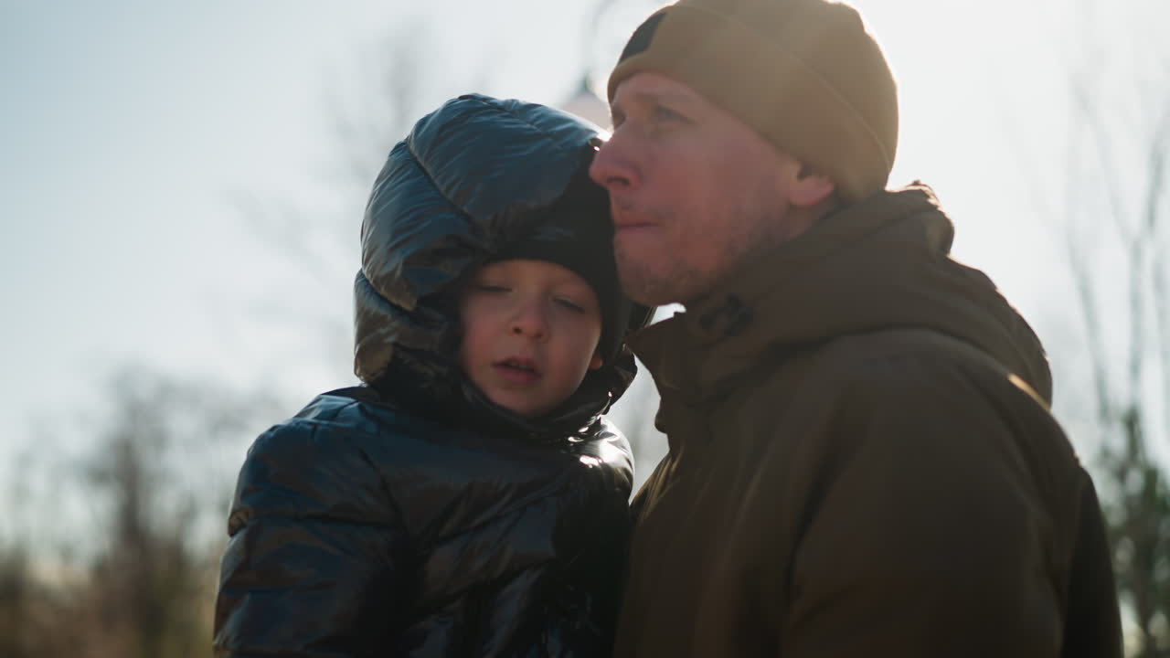 Father carrying his son and speaking with the child, both are dressed in jeans and with the child wearing a puffy jacket and the man in a beanie, with electric poles behind