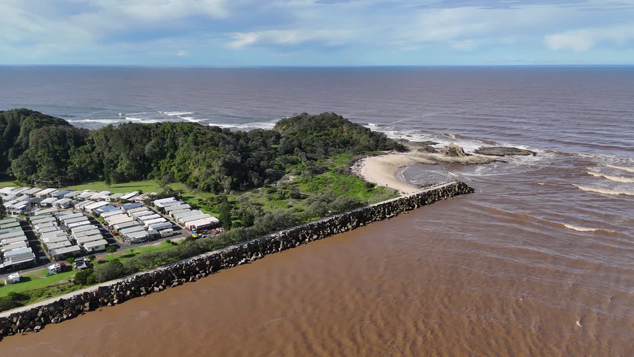 Aerial drone footage over riverside caravan park, brown river, lush greenery, sandy beach, ocean