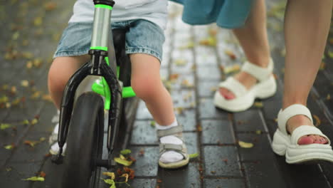 close-up of a child on a bicycle with a woman walking closely with him on a leaf-covered pathway, the child drops his leg for balance as he maneuvers through the pathway