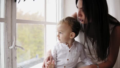 la joven madre está sentada con su hijo en el alféizar de la ventana decorada con una corona de navidad y mirando hacia afuera. ellos están hablando