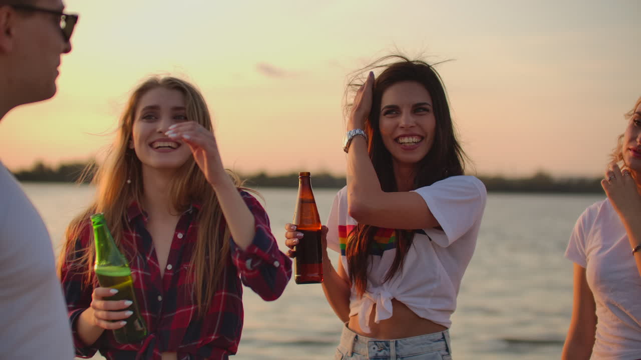 Two female students are touching their long hair and talking with a friend on the beach party with beer. A great summer open air party next to the lake at sunset.