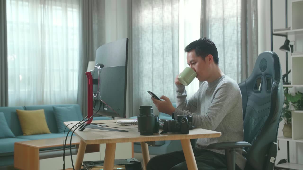 Asian Cameraman In Long Sleeved T-Shirt And Black Pants Using Mobile Phone And Drinking Coffee While Using Desktop Computer For Working At Home.