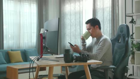 asian cameraman in long sleeved t-shirt and black pants using mobile phone and drinking coffee while using desktop computer for working at home.