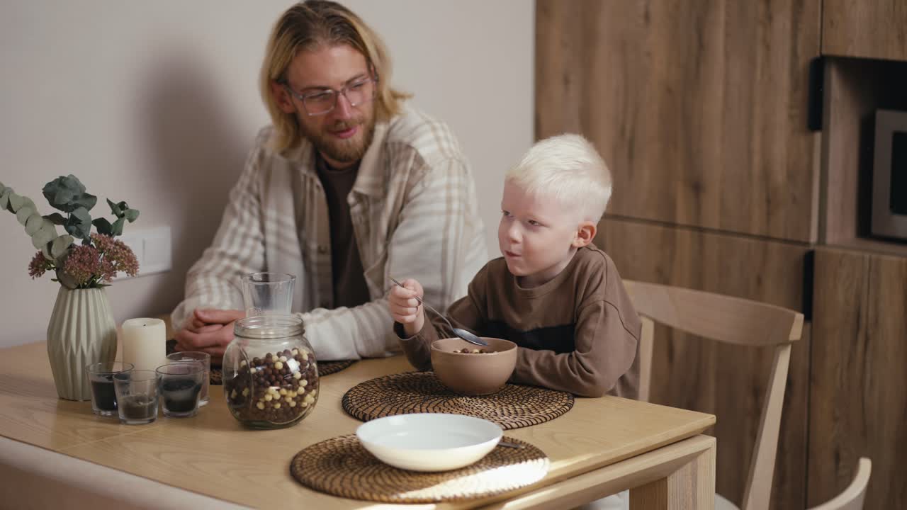 Happy little albino boy with white hair color in a brown jacket eats cereal with milk and has breakfast with his blond father with a beard and glasses in a modern kitchen in the morning