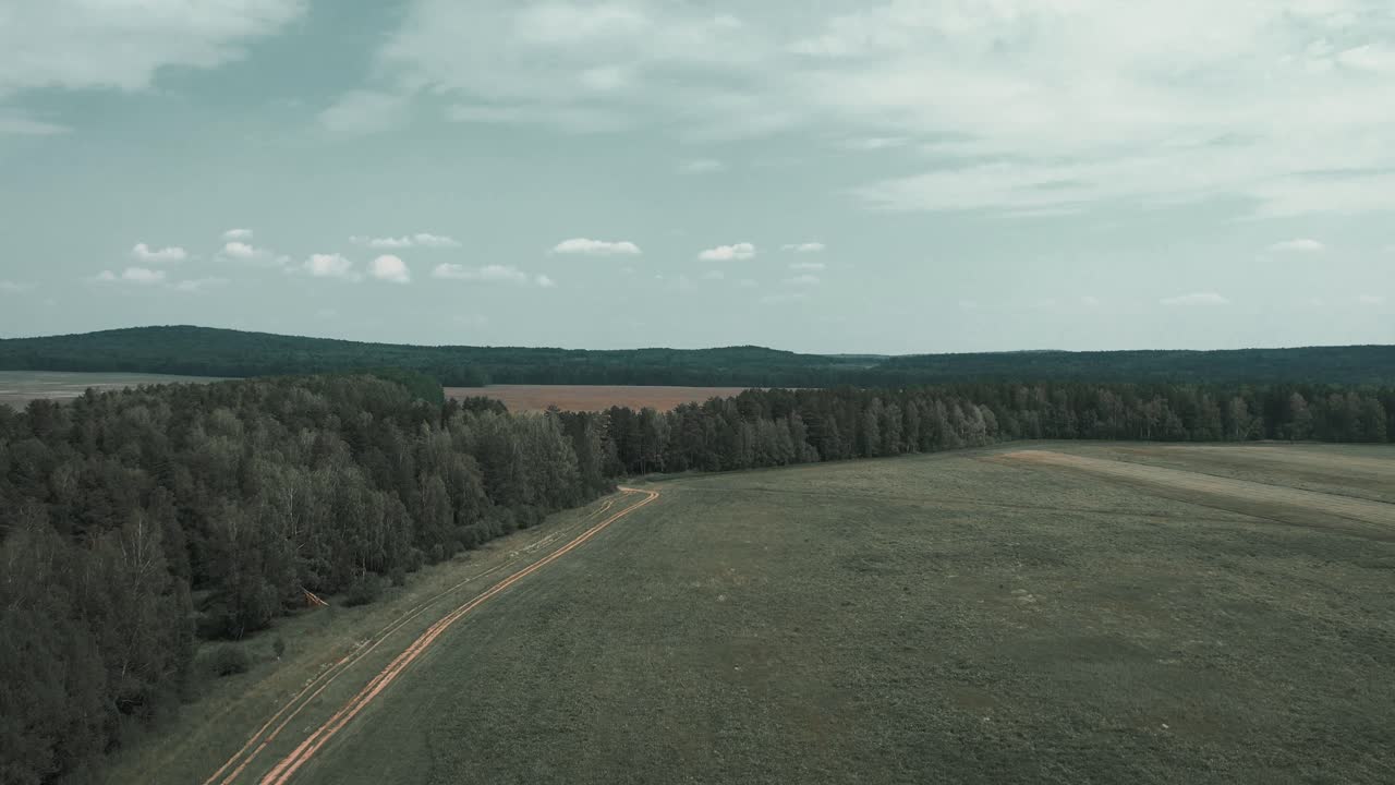 Rusya, Ural Dağları bölgesinde, orman kenarı boyunca uzanan virajlı bir toprak yolun ve bulutlu gökyüzü altındaki açık alanların drone görüntüsü, sakin kırsal manzara.