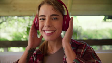 Blonde-girl-in-a-red-plaid-shirt-puts-on-red-wireless-headphones-smiles-looks-at-the-camera-and-shakes-her-head-Sitting-on-a-sofa-in-a-gazebo-in-nature