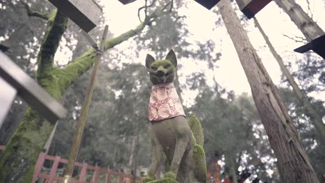 View-Of-Fox-Statue-With-Scarf-And-Tree-Branches-Covered-With-Moss-In-Fushimi-Inari-Shinto-Shrine-In-Kyoto,-Japan---Low-Angle-Shot