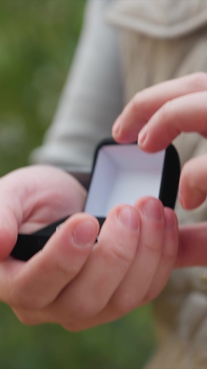 Tessuto di capospalla beige con dettagli di cucitura visibili. La mano sinistra afferra il bordo inferiore di un oggetto rettangolare scuro. Una ripresa fissa si concentra sull'interazione con un piccolo oggetto.