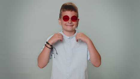 seductor alegre niño pequeño elegante niño en camisa con gafas de sol sonrisa encantadora en la pared azul