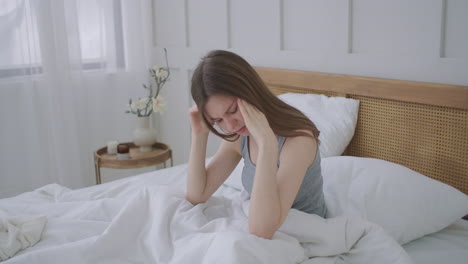 women sitting on bed holding her head. she has a painful headache. the woman experiences a headache rubs her temples and frowns in pain. seasonal disease caucasian girl in the bedroom on sick leave