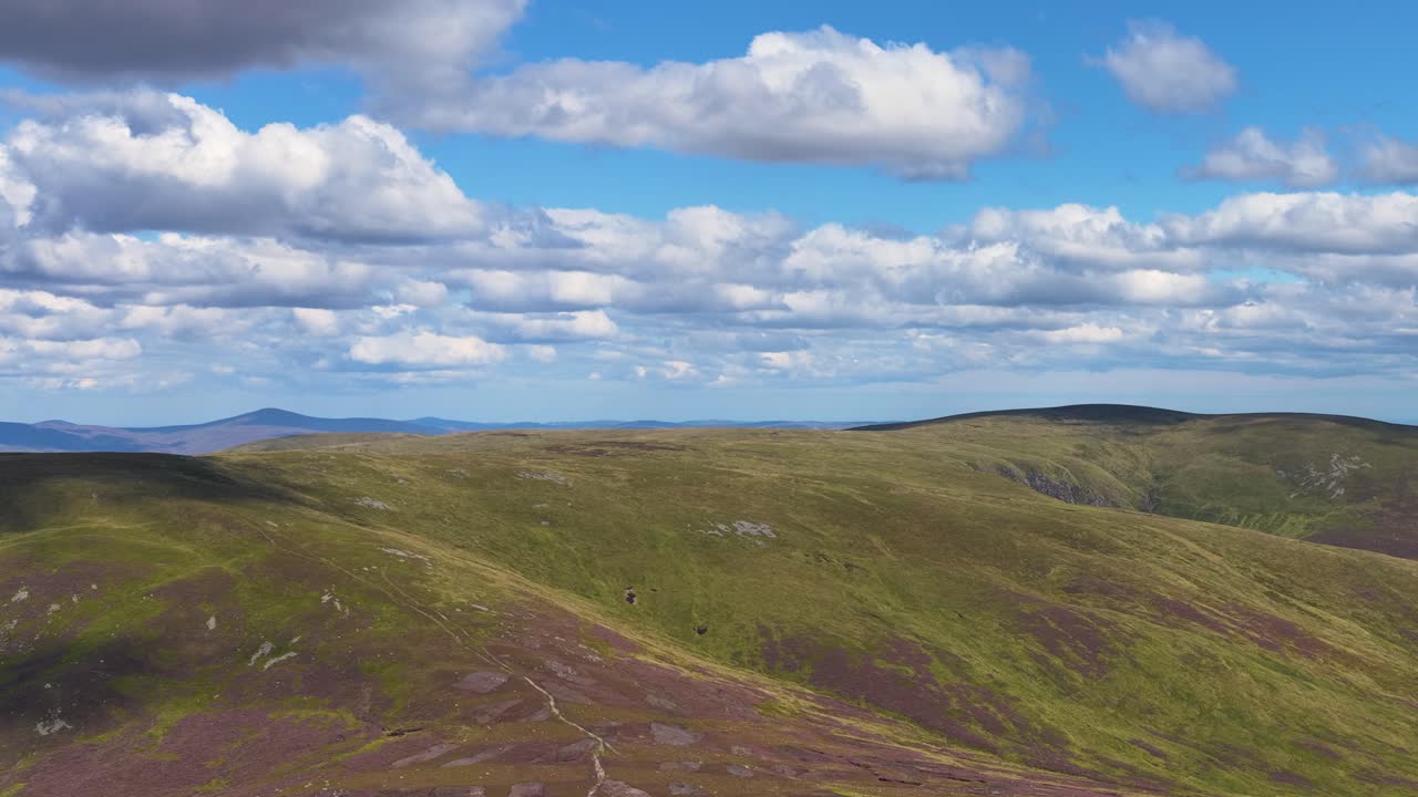 Drone pans across tranquil Scottish Highlands, revealing rolling heather hills, blue sky, and scattered clouds