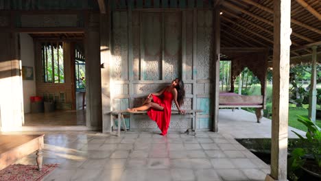 young asian female posing on wooden bench in joglo style bali villa with a beautiful red maxi dress