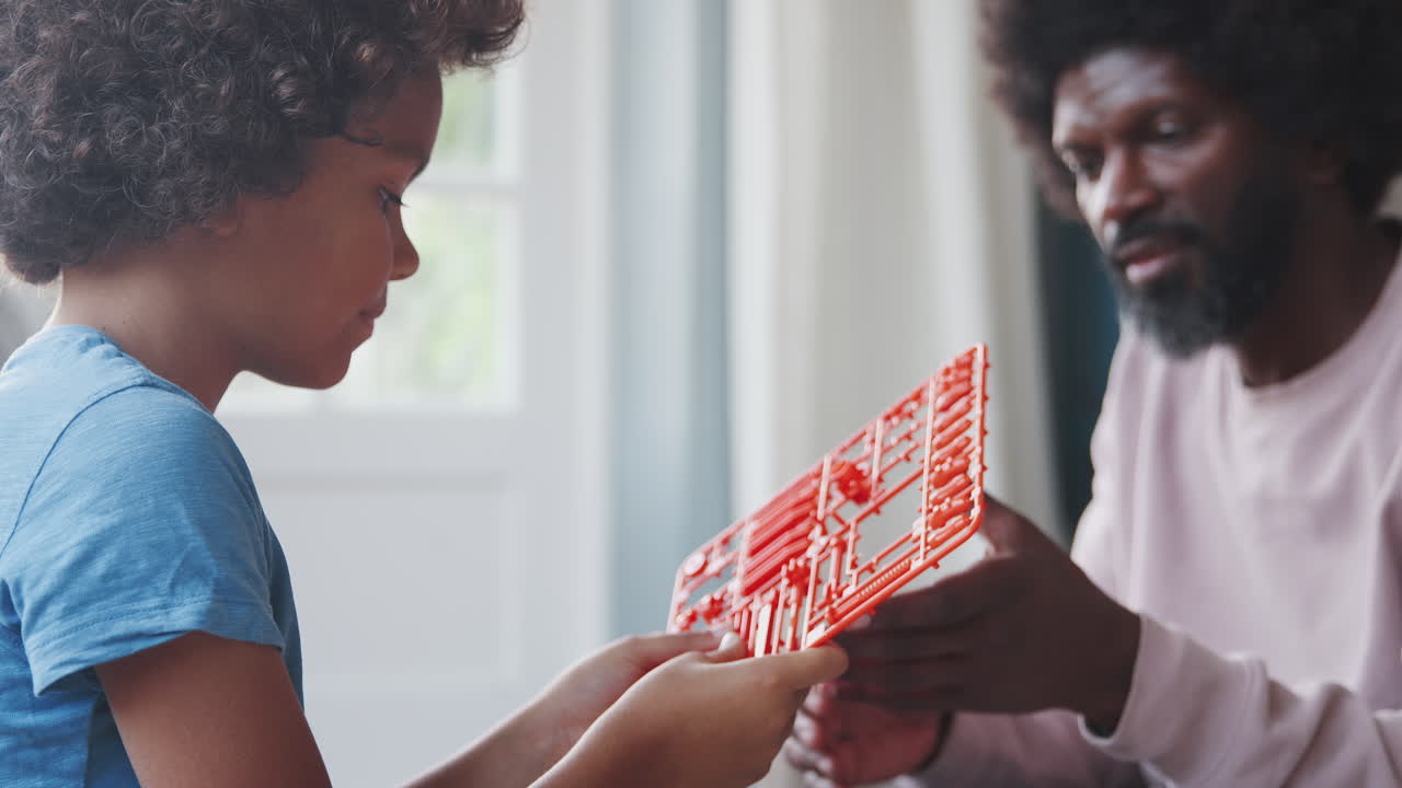 padre negro de mediana edad sentado en el suelo en casa ayudando a su hijo pre-adolescente a separar las piezas de un kit de construcción de juguete, de cerca, enfoque selectivo