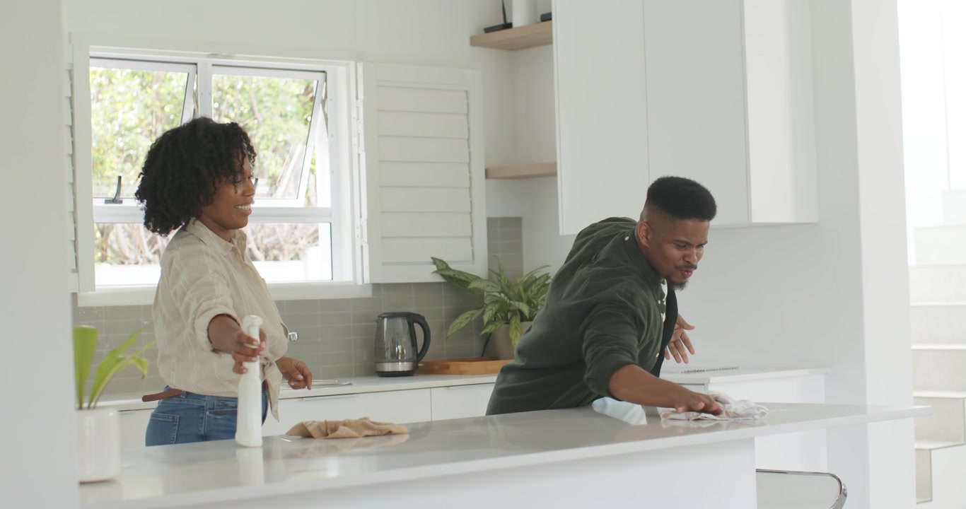 Coppia afroamericana che pulisce il bancone in cucina, la donna che porge lo spruzzino mentre l'uomo pulisce con un asciugamano.