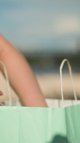 primer plano de una mujer sentada en un banco al aire libre con bolsas de compras a ambos lados, recupera un libro de una bolsa verde menta, lo abre y hojea las páginas, con un fondo borroso