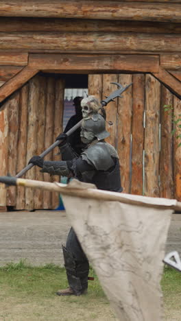 duel between dark knight in armor and young female near wooden log building. warriors fight on spears skillfully standing in courtyard of medieval village