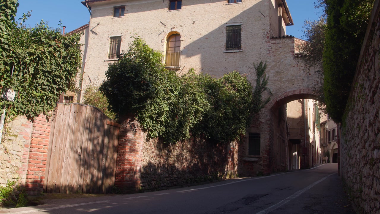 Strada tranquilla che passa sotto un arco storico in pietra con vecchi edifici e vegetazione.