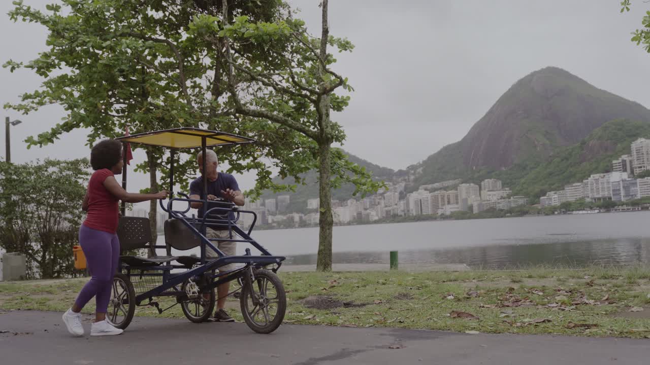 Two people with a pedal bike by a lake with a mountain in the background