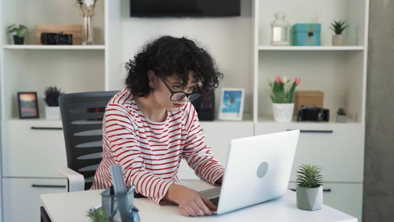 The curly-haired young woman is sitting at a desk with a laptop in a nice bright office, putting on glasses for working on the laptop