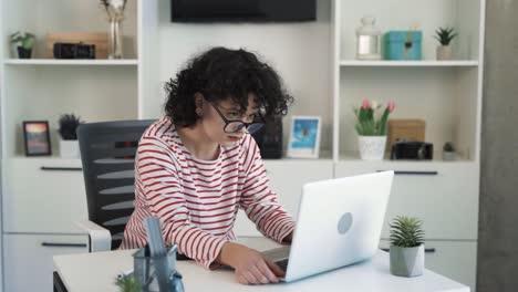 the curly-haired young woman is sitting at a desk with a laptop in a nice bright office, putting on glasses for working on the laptop