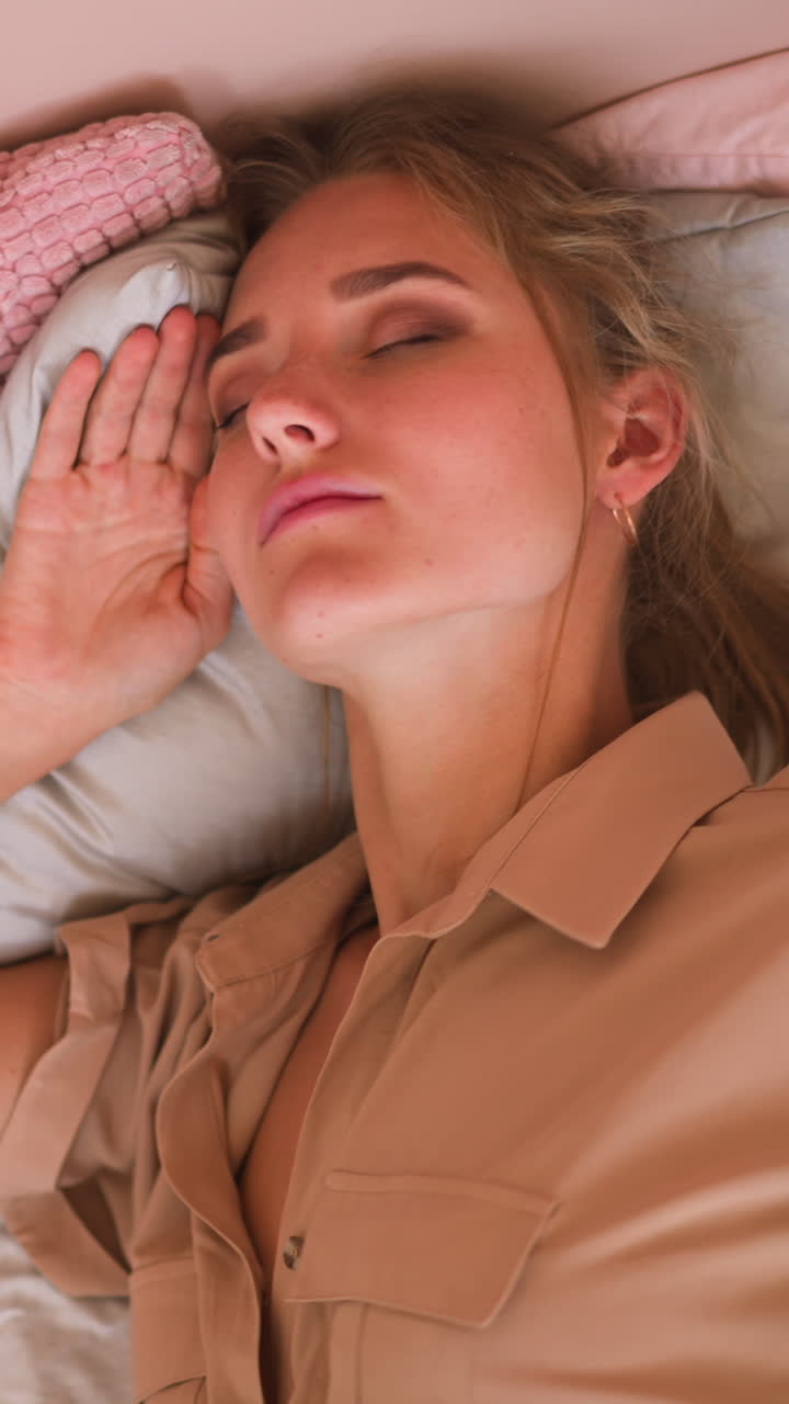 Tired woman with fair hair turns on cozy bed and covers head with soft white pillow trying to sleep after hard working day at home slow motion