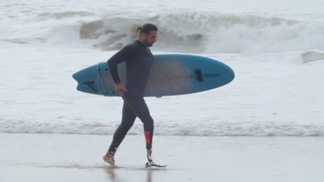 tracking shot of a disabled man in wetsuit with artificial leg running along coastline with surfboard 1