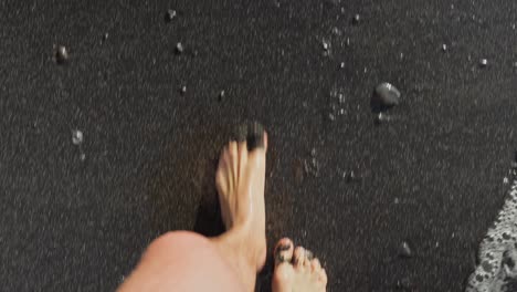 Top-down-POV-of-person-walking-along-black-pebble-sand-beach-as-ocean-waves-recede