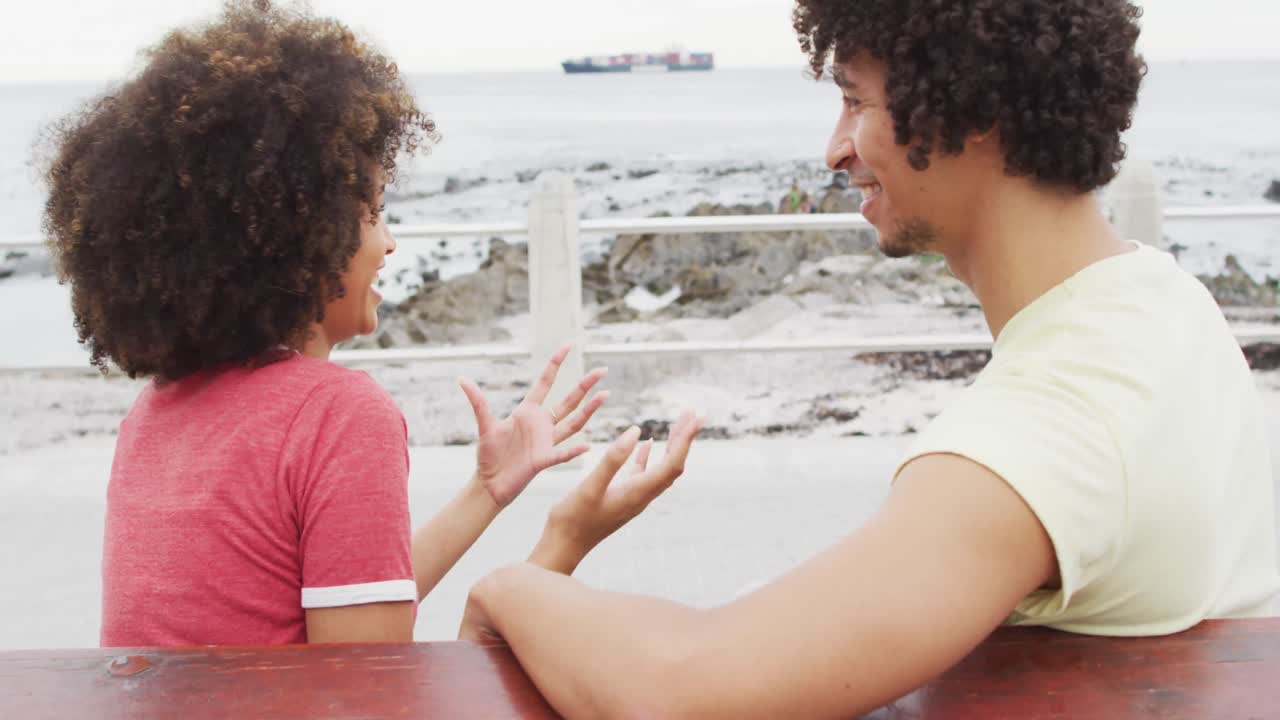 pareja afroamericana sonriendo mientras hablan entre sí sentados en un banco en el paseo marítimo